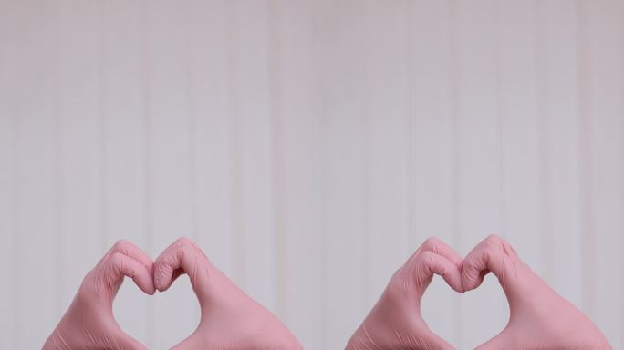 垂直视频的人在粉红色乳胶手套显示心脏手势对白色背景，近距离对手。双手戴上心形手套，象征对医疗队的爱和