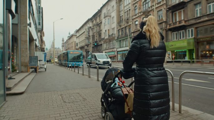匈牙利布达佩斯市，一名身穿冬衣的女子推着婴儿车走在人行道上的后视图