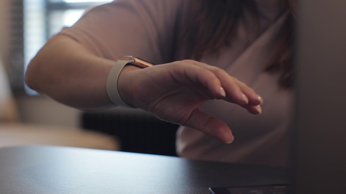 女性手指可以双击智能手表，实现免提交互。年轻女子正在办公室的工作桌旁用笔记本电脑打字。