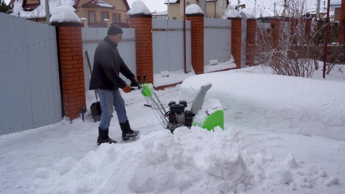 冬天，一个人在房子的院子里打扫雪，一个人用吹雪机打扫雪