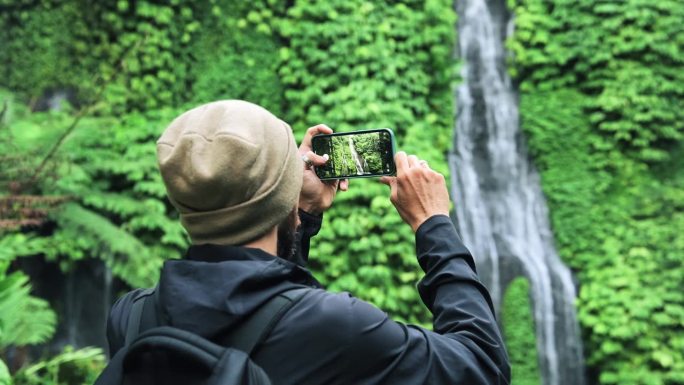 在绿色大自然中旅行的成年男子用手机拍下了森林瀑布。在丛林瀑布旅行目的地的年轻人的生活方式。旅行者制作