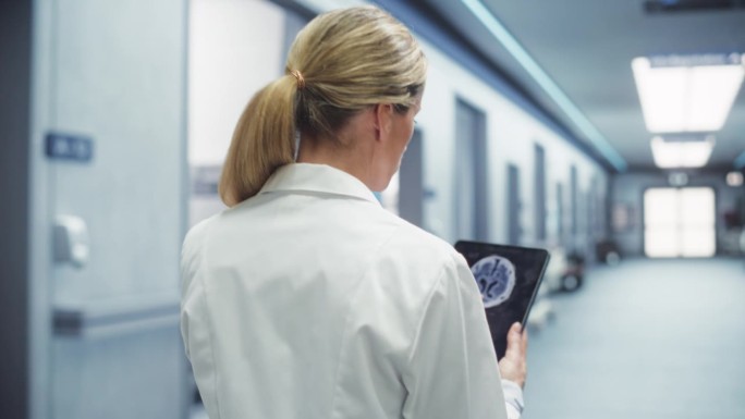 女医生用平板电脑在医院走廊行走的后视跟踪镜头。资深女外科医生在手术前检查脑核磁共振成像，重温她的笔记