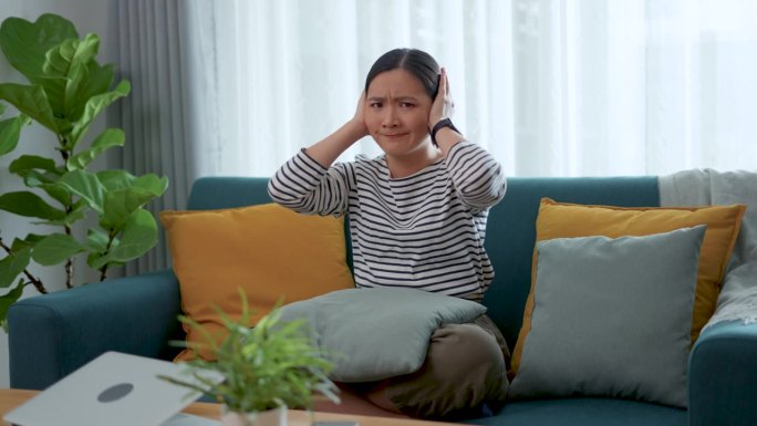亚洲女性坐在家里捂着耳朵，对噪音感到厌烦。