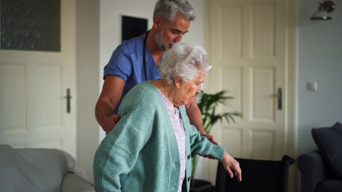 护工帮助家中的老年妇女从轮椅上站起来。