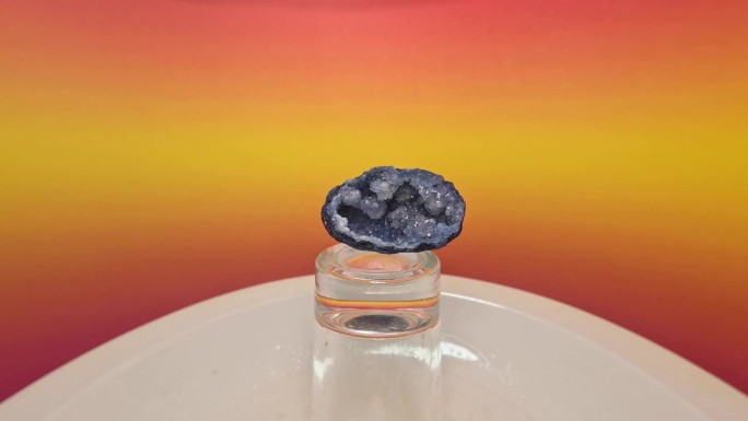 蓝色晶洞水晶石在一块显示玻璃柱上缓慢旋转，背景模糊，色彩斑斓