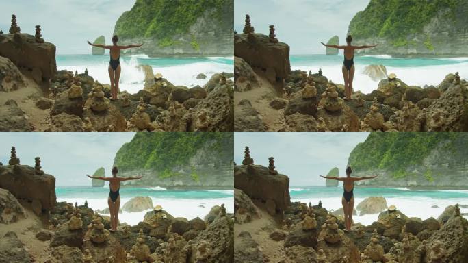 穿着黑色泳装的瘦骨嶙峋的年轻女子在海浪附近的海滩上张开双臂。女孩在岩石岸边放松。夏天休息。后视镜，后