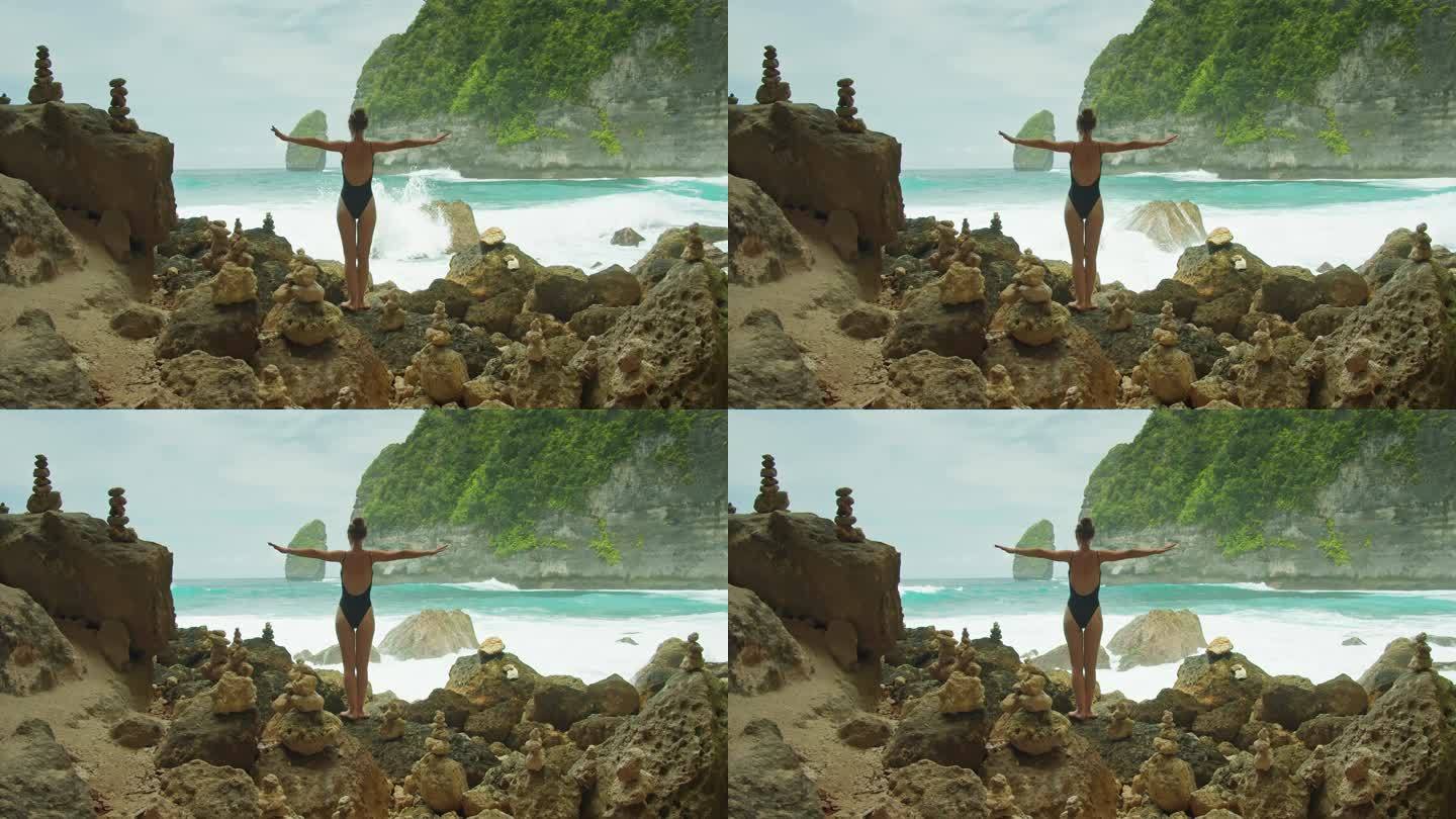 穿着黑色泳装的瘦骨嶙峋的年轻女子在海浪附近的海滩上张开双臂。女孩在岩石岸边放松。夏天休息。后视镜，后