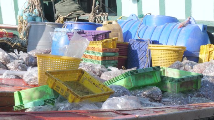一艘上下浮动的渔船停靠在渔港，甲板上堆满了装着鱼的塑料袋、板条箱和篮子