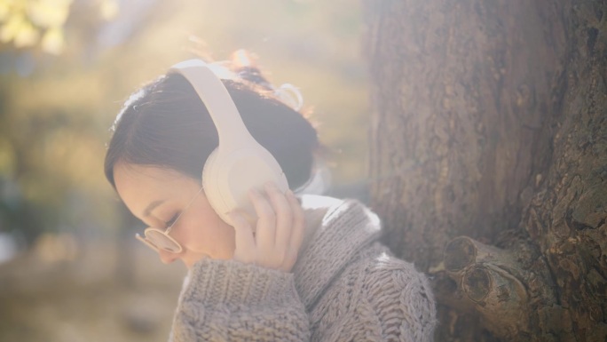 一位孕妇戴着白色耳机听音乐。秋天，她站在公园的一棵树下。