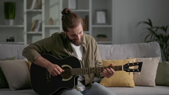一个留着长发和胡子的男人坐在沙发上弹吉他。