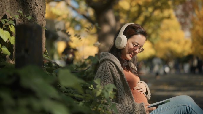 孕妇坐在公园里，在树下放松，看着落叶，听音乐。