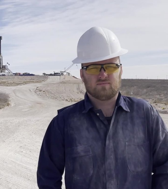年轻的油田水力压裂钻井工人在钻井现场前，穿着全套工作服和安全帽，看着相机和折叠手臂