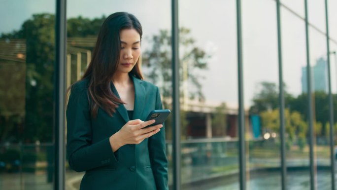 年轻漂亮的亚洲女商人使用手机，玩社交媒体，通过在线消息与人聊天，在休息时间站在户外办公室，商务人士与