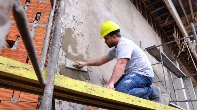 一名蓝领工人跪在脚手架上，在建筑工地给墙浇水泥