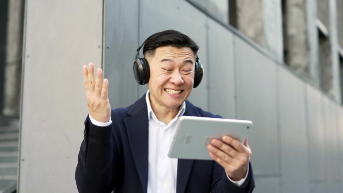 兴奋的亚洲商人穿着正装，戴着无线耳机，用数码平板电脑观看体育比赛或比赛
