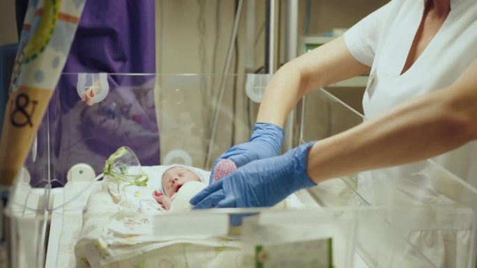 不知名的护士戴着白手套，采取行动照顾早产儿，选择性地关注婴儿的眼睛。新生儿放在保温箱中。新生儿重症监
