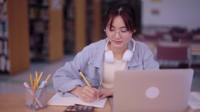 专注的亚洲女学生进行研究，收集信息，并在大学图书馆的笔记本上记录调查结果。在学术环境中孜孜不倦地追求