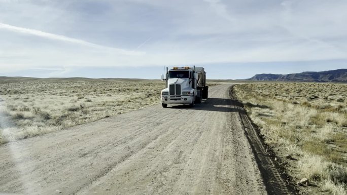 在一个阳光明媚的冬日，手持拍摄的一辆白色半挂车在犹他州沙漠的土路上拖着一辆自卸拖车