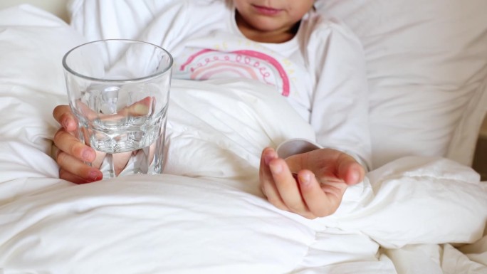 特写:小孩双手掌心握着玻璃杯、水和药片。生病的孩子躺在床上准备吃药、补充维生素、矿物质、营养、保健、