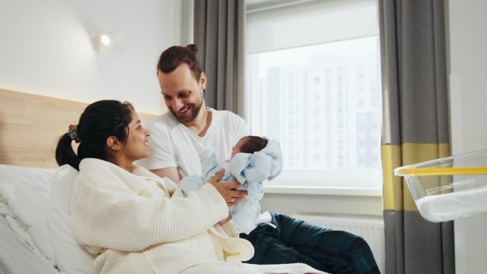 幸福的父母抱着刚出生的男婴在医院病房。微笑的母亲坐在病床上，丈夫在身边，抱着婴儿。父亲将襁褓中的新生