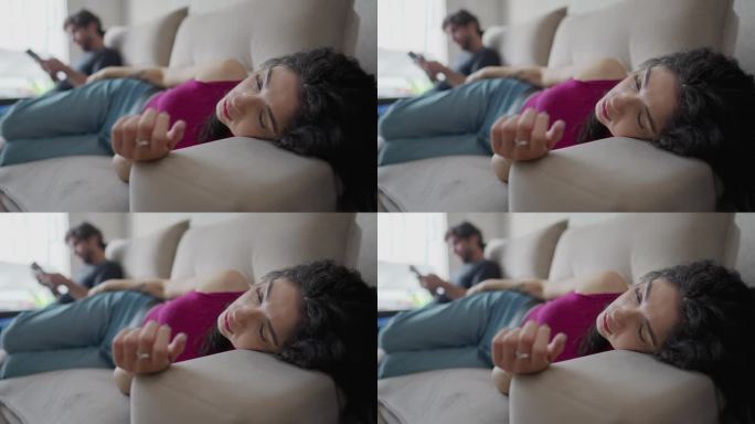 疲惫的年轻女子躺在沙发上，脸上带着疲惫的表情，而男友在后台看手机。孤独的人独自在一起