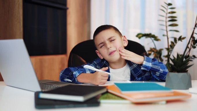 悲伤的小学生闭着眼睛摸着脸颊，在家里学习时牙疼。穿着便服的抑郁男孩有疾病症状。