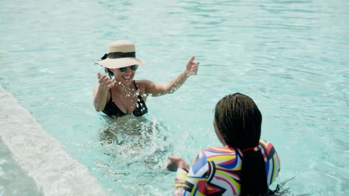两个非洲裔，西班牙裔妇女彼此是外国人，但两个朋友，一起在游泳池里玩，在早上玩得很开心，他们用手开心地