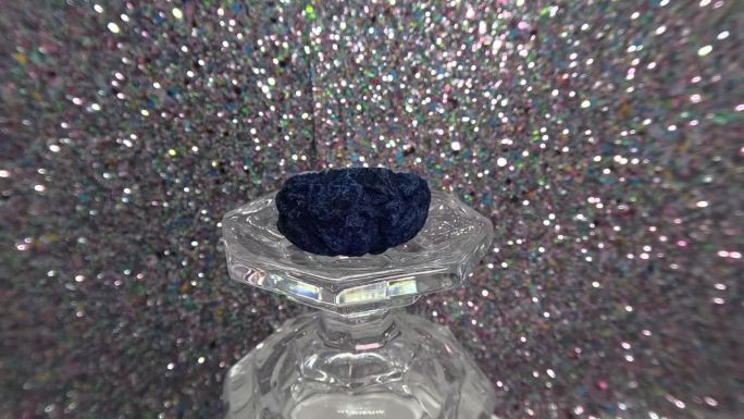 蓝色晶洞水晶在翻转的玻璃杯上缓慢旋转，背景是模糊的银色闪光