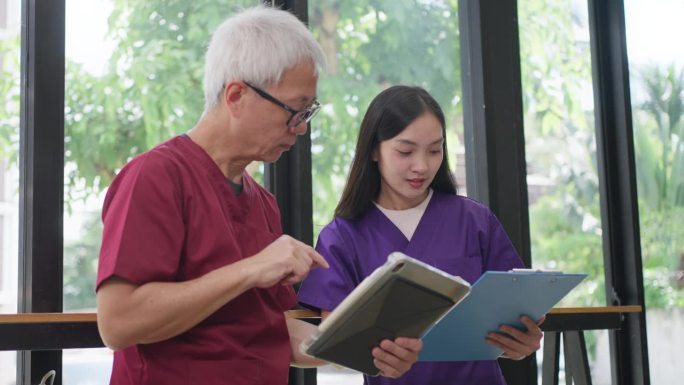 穿着手术服的亚洲女护士一边讲笔记，一边和一位年长的男医生交谈，这位男医生手里拿着一台平板电脑，向她展