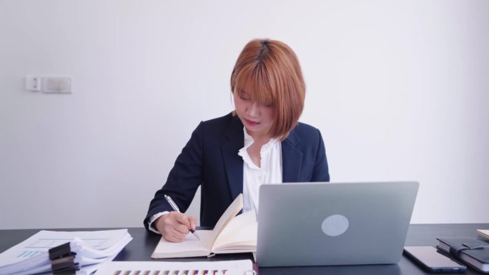 一位自信的亚洲女商人在她专门的工作空间里，孜孜不倦地把公司的重要数据从笔记本电脑记录到记事本上