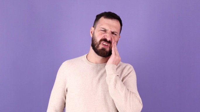 有牙齿疼痛问题的大胡子男子在工作室的紫色背景中被隔离。