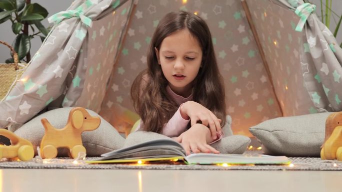好奇的黑发小女孩坐在家里的棚屋地板上读纸质书翻页看图片，享受新教科书的故事。