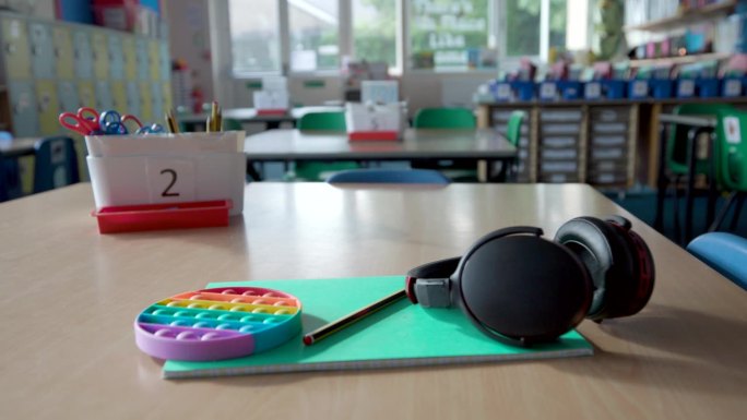 在空荡荡的学校教室里，桌子上放着护耳器、耳机和烦躁玩具等帮助自闭症儿童的工具