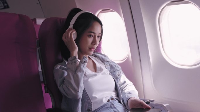 目前，乘坐飞机旅行的亚洲女性正愉快地戴着耳机听音乐，轻声哼唱。能唤起快乐和放松。