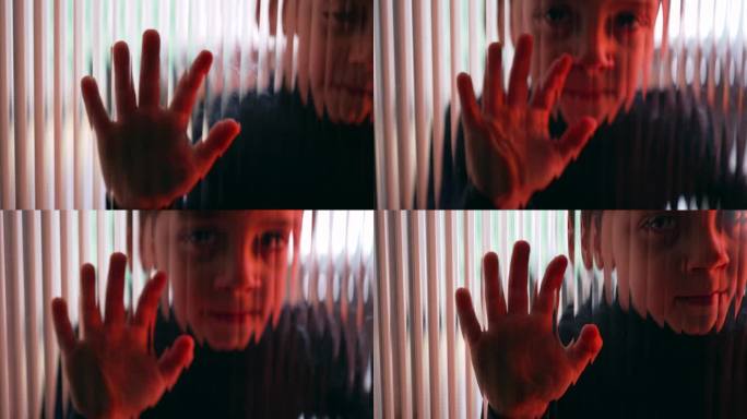 一个小男孩靠在散焦的红色窗户上。孩子隔着玻璃看相机