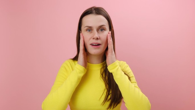 25岁的年轻女性穿着黄色的毛衣，在摄影棚的粉红色背景墙上独自摆姿势，惊讶地张大嘴巴，竖起大拇指，表现