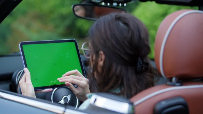 坐在车里滚动绿屏平板电脑的女人。女司机在车内使用色度键装置。从肩膀上看。