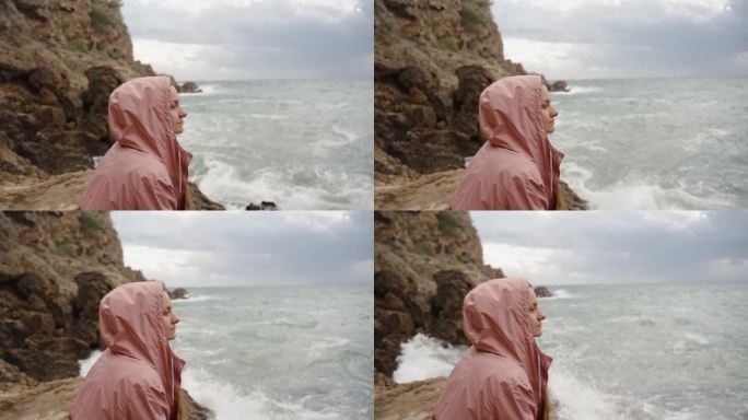 一位身穿粉色外套的年轻女子凝视远方，陷入沉思。当她坐在岸边时，大浪冲击着岩石。缓慢的运动。