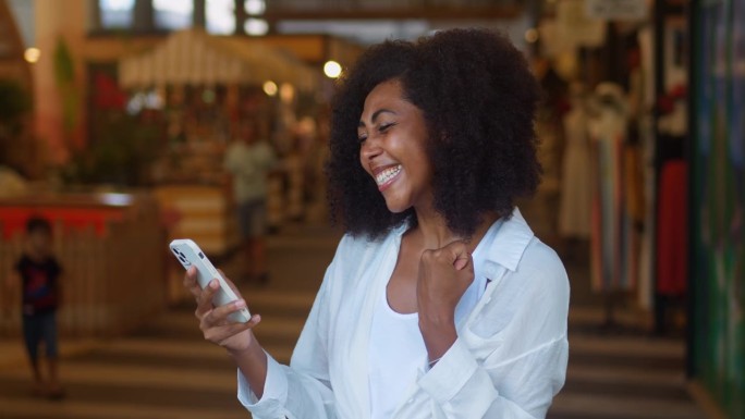 一名身穿白色休闲服装的非裔美国女性站在商场内，一边用智能手机接收好消息，一边咧着嘴笑，握紧拳头，跳着