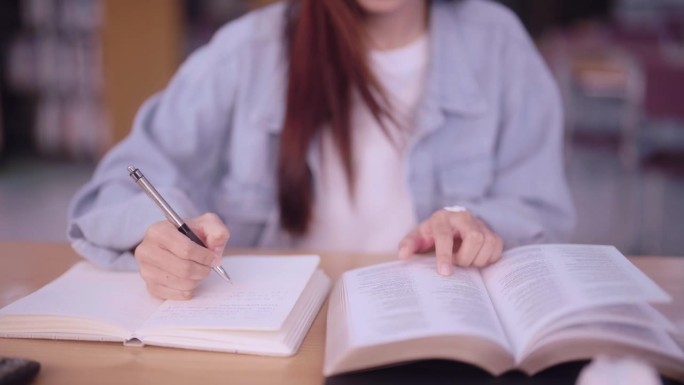 勤奋的亚洲女学生在她的大学图书馆的书上做笔记，展示了一种致力于学习和利用物质资源取得学术成就的方法。