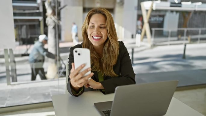 微笑的西班牙女性在现代办公室室内使用智能手机，展示科技，专业和生活方式的元素。