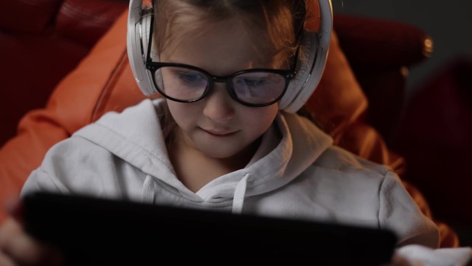 兴奋的少女坐在家里的沙发上，用数字平板电脑技术设备玩游戏。情绪化的孩子戴着耳机和眼镜，拿着pad电脑
