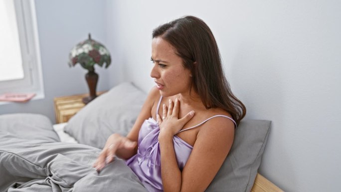 一位体弱多病但美丽的西班牙年轻女子穿着睡衣坐在卧室的床上，因支气管炎或感冒而咳嗽，这是医疗保健之战的