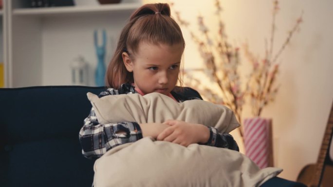 心烦意乱的小女孩独自坐在沙发上，抱着枕头，不开心的孩子
