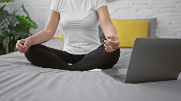 一名女子醒着，注意力集中，在做平静的瑜伽练习，双手休息，坐在床上，笔记本电脑旁边的在线指导，让她的卧