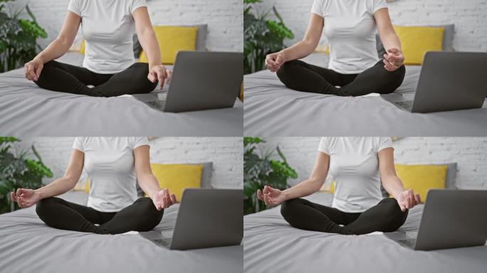 一名女子醒着，注意力集中，在做平静的瑜伽练习，双手休息，坐在床上，笔记本电脑旁边的在线指导，让她的卧