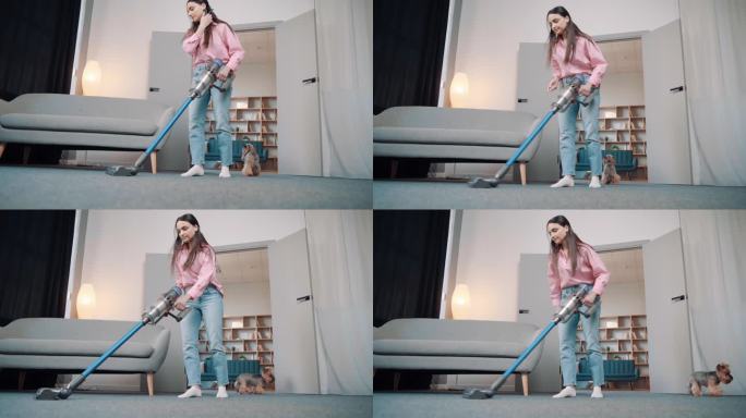 一个年轻的欧洲女孩穿着舒适的家居服，用无线吸尘器在公寓里清理地毯上一只金色约克夏梗犬的狗毛