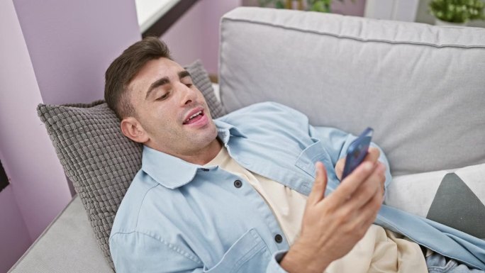 开朗的年轻西班牙人躺在家里的沙发上，全神贯注地用智能手机发短信，在舒适的客厅里散发着积极的光芒。