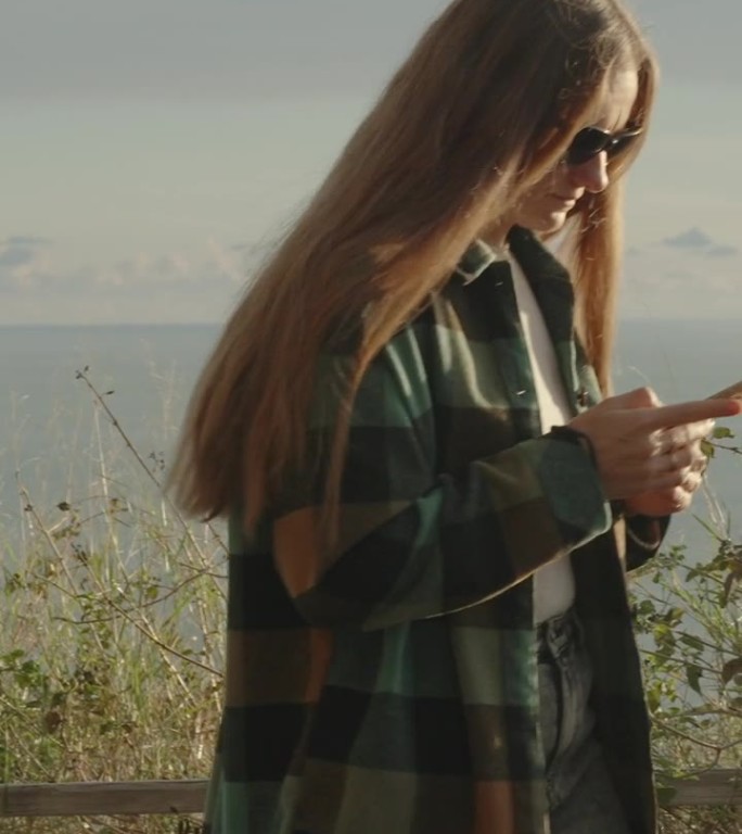 一位年轻的长发女子走在高山的海边，目不转睛地看着她的手机。日落。