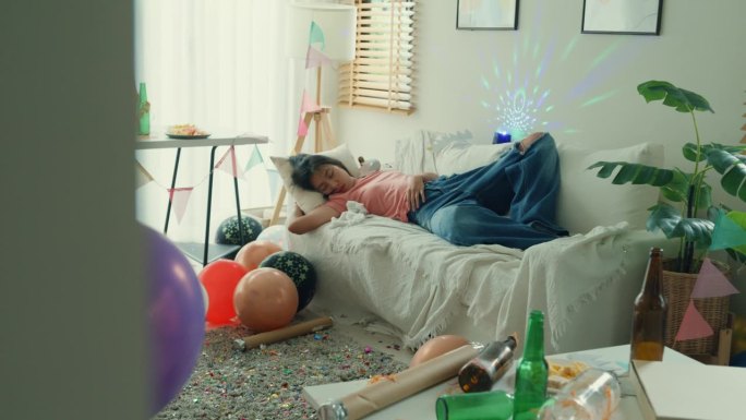 醉酒的年轻亚洲女子在家里庆祝派对后睡在沙发上。疯狂的家庭聚会后凌乱的房间。
